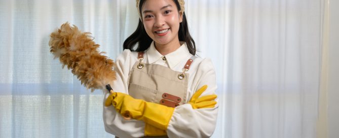 Pembantu rumah muda Asia memakai apron dan sarung tangan kuning, tersenyum cerah sambil memegang kain penyapu bulu, bersedia untuk menangani tugas pembersihan rumah dengan penuh semangat