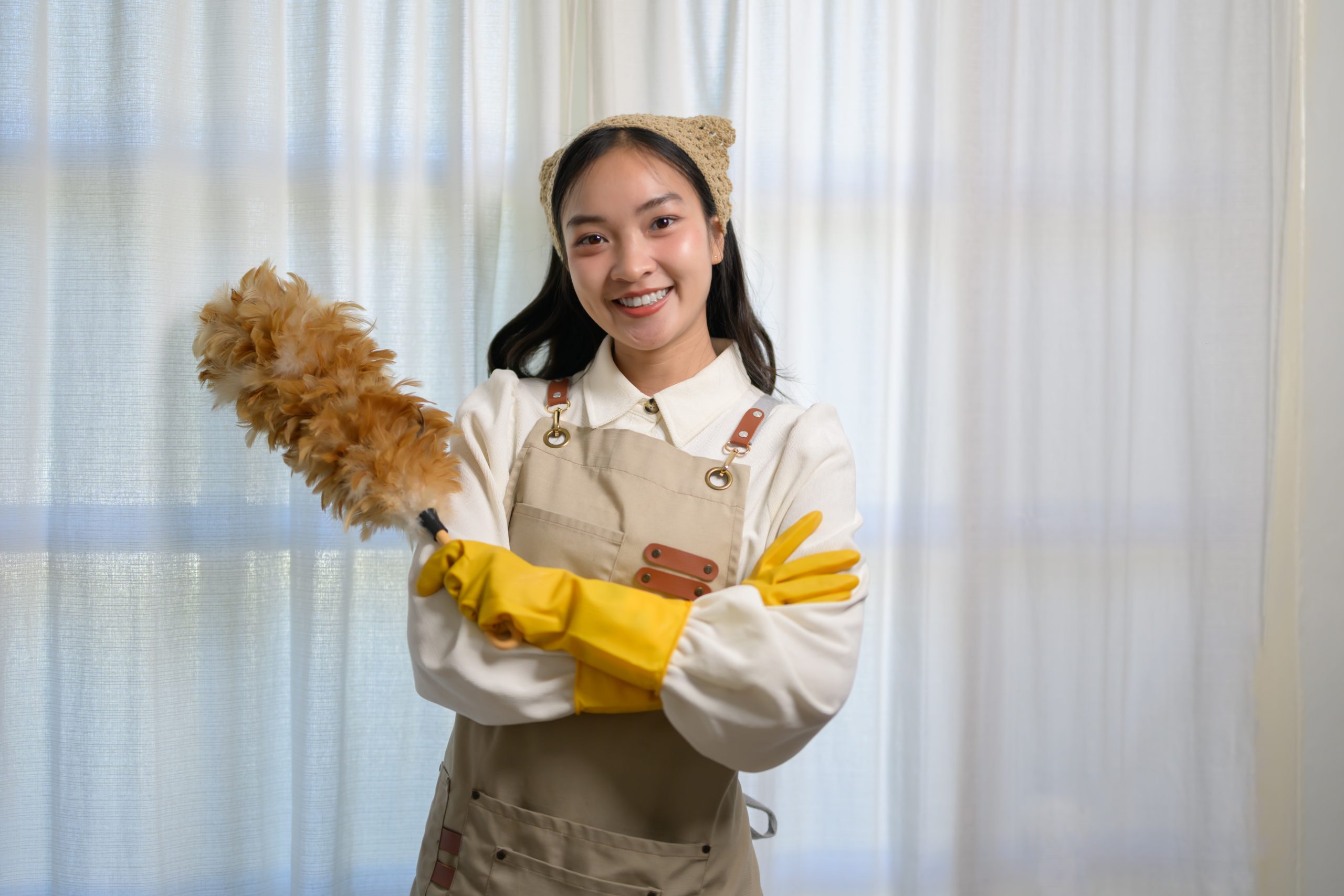 Pembantu rumah muda Asia memakai apron dan sarung tangan kuning, tersenyum cerah sambil memegang kain penyapu bulu, bersedia untuk menangani tugas pembersihan rumah dengan penuh semangat